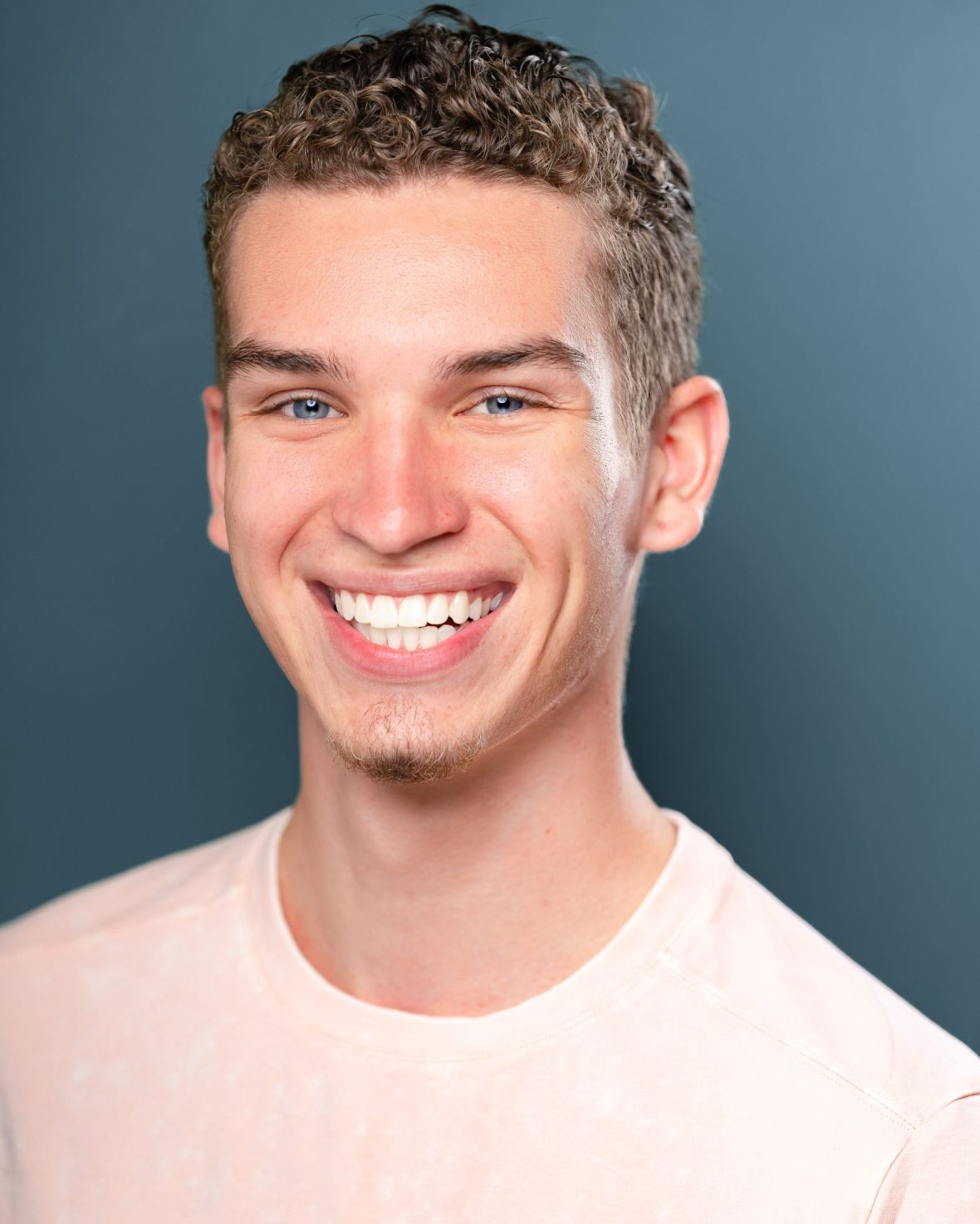 Headshot of theatre student with a bright smile wearing a pink shirt against a grey backdrop