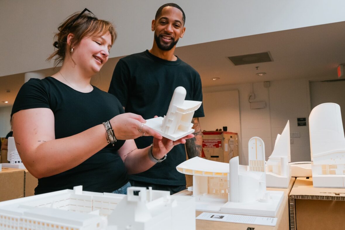 Two individuals stand before a detailed model of a building, engaged in discussion about its design and features.