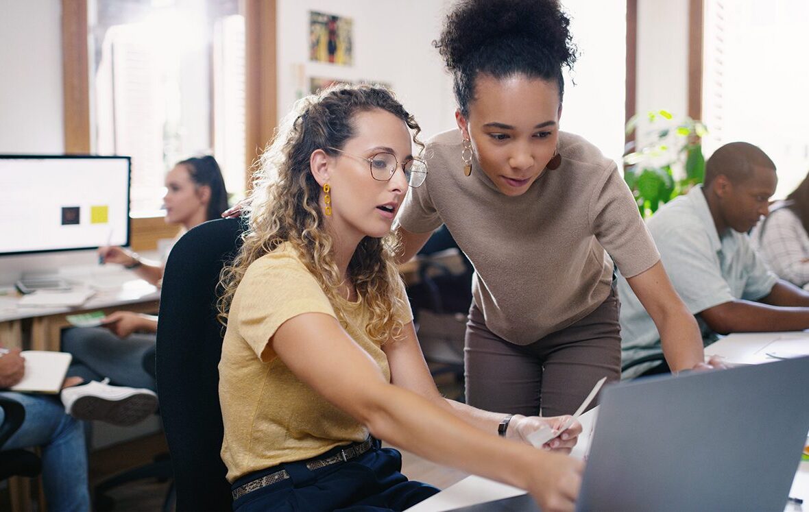 Two women collaborate on a laptop in a busy office.