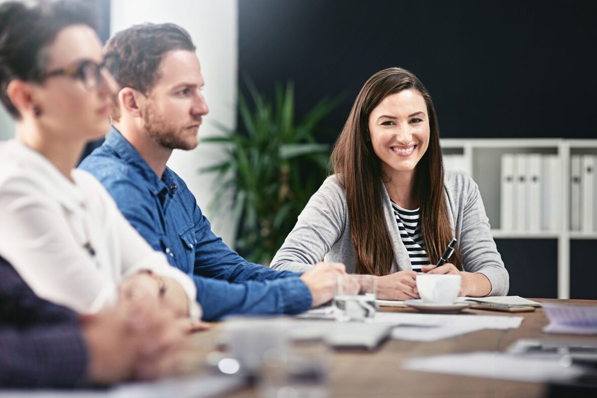 A woman smiling at a conference table with two colleagues in a professional setting.